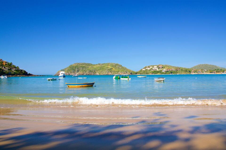 A beach with clear blue water, boats, and hills in the background.