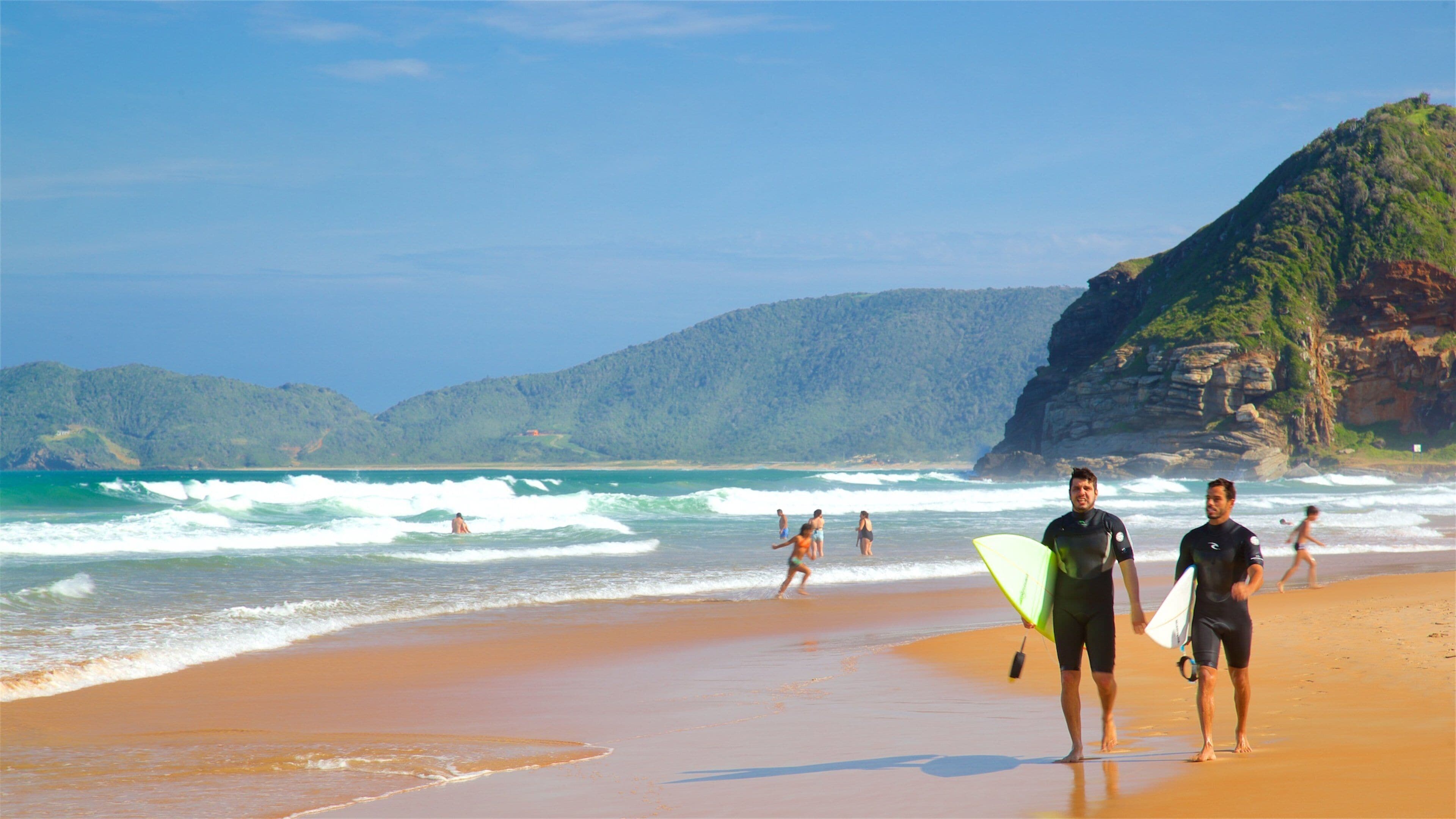 A beach with surfers walking along the shore, waves crashing, and a mountainous backdrop.