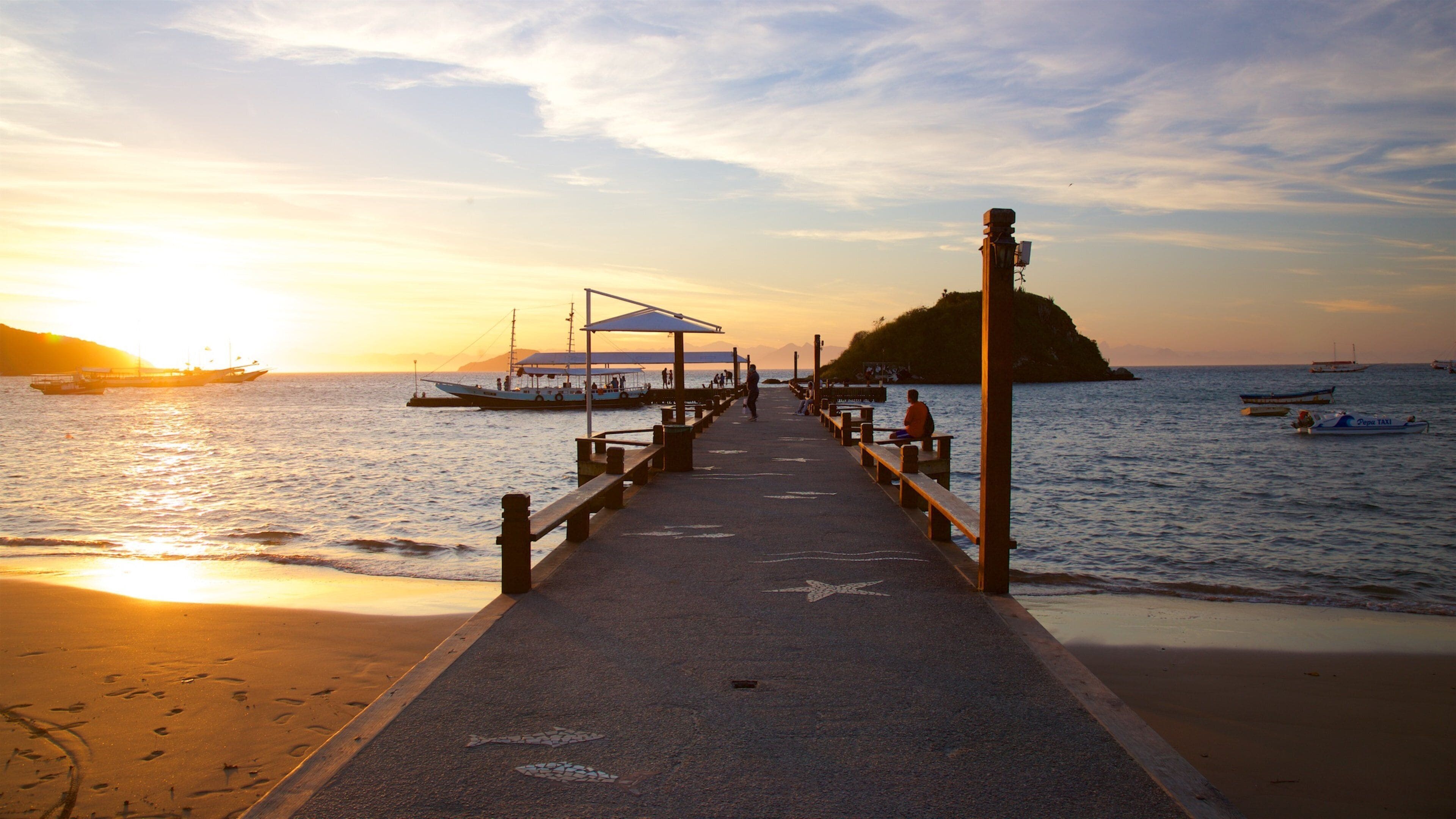 A pier extending into calm waters at sunset with boats and a small island in the background.