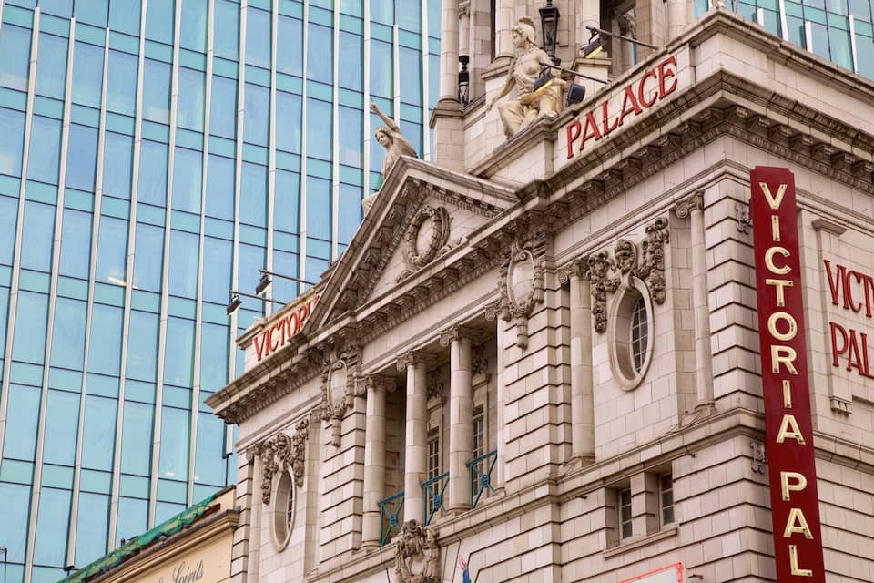Exterior do Teatro Victoria Palace, com detalhes arquitetônicos ornamentados e um edifício moderno de vidro ao fundo.