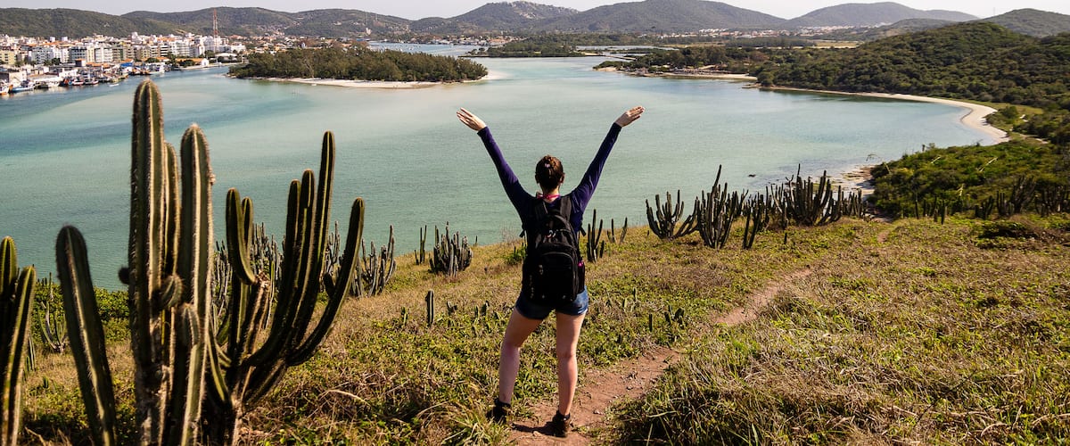 A person standing on a path with arms raised, overlooking a coastal landscape with cacti, a beach, and a distant city.