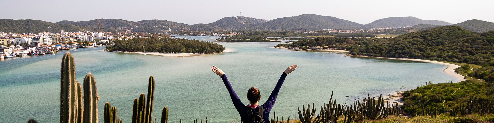 A person standing on a path with arms raised, overlooking a coastal landscape with cacti, a beach, and a distant city.