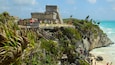 A coastal archaeological site with a large stone structure, wooden walkways, and visitors.