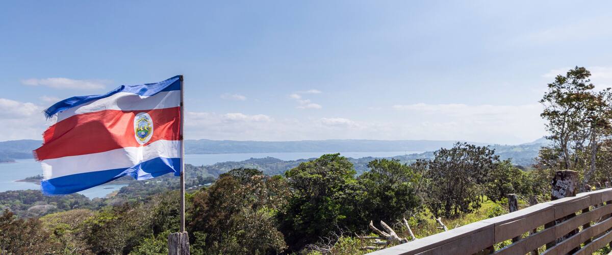 A flag on a pole, a wooden fence, and a landscape with trees and a body of water in the distance.
