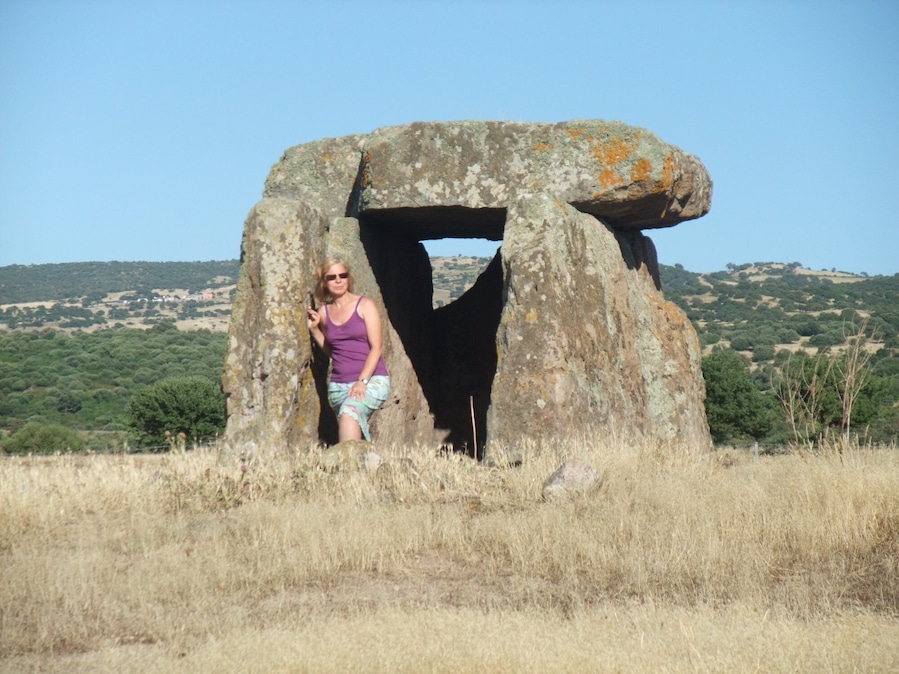 Una persona de pie frente a una gran estructura de piedra en un campo cubierto de hierba, con colinas al fondo.