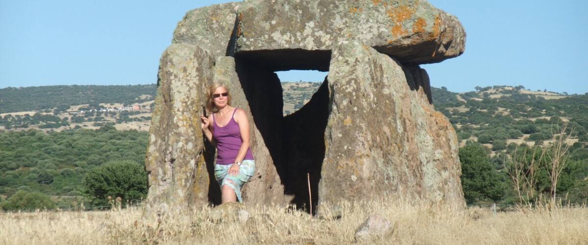 A person standing in front of a large stone structure in a grassy field with hills in the background.
