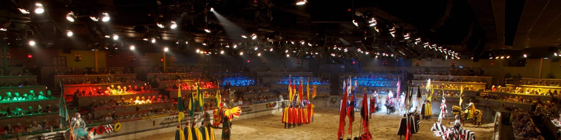 A medieval jousting tournament in a large indoor arena with sand flooring and spectators in the stands.