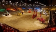 A medieval jousting tournament in a large indoor arena with sand flooring and spectators in the stands.