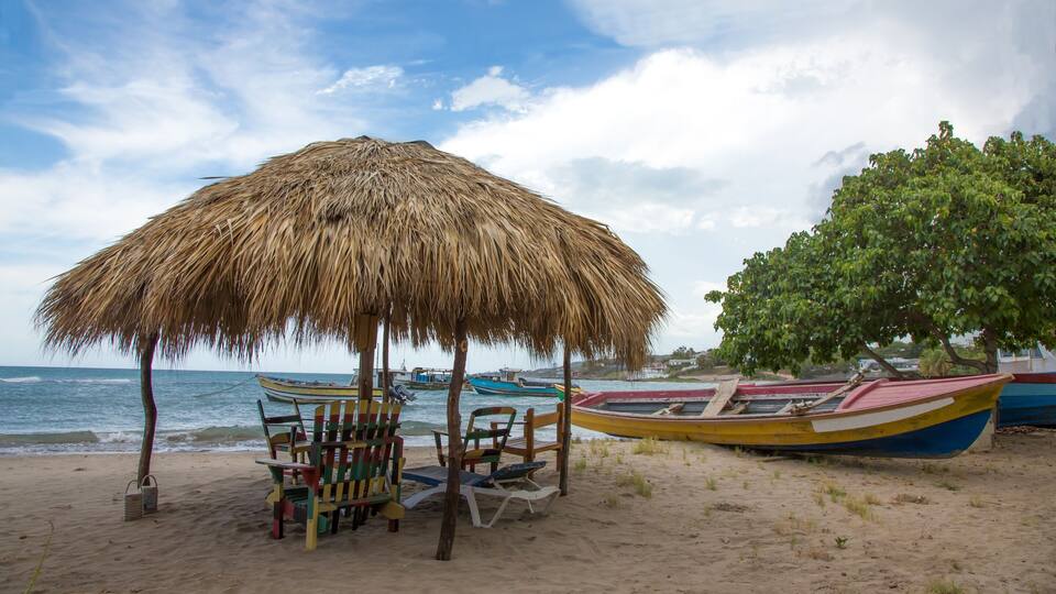 A beach with a thatched umbrella, colorful chairs, and boats.