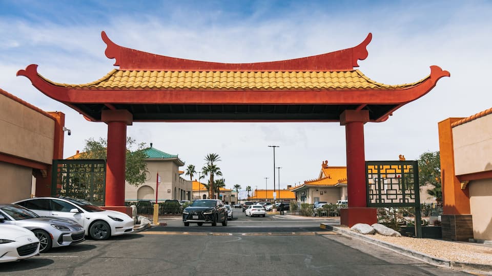 A traditional Chinese gate with red pillars and yellow tiles, leading to a street lined with parked cars and buildings with orange roofs.