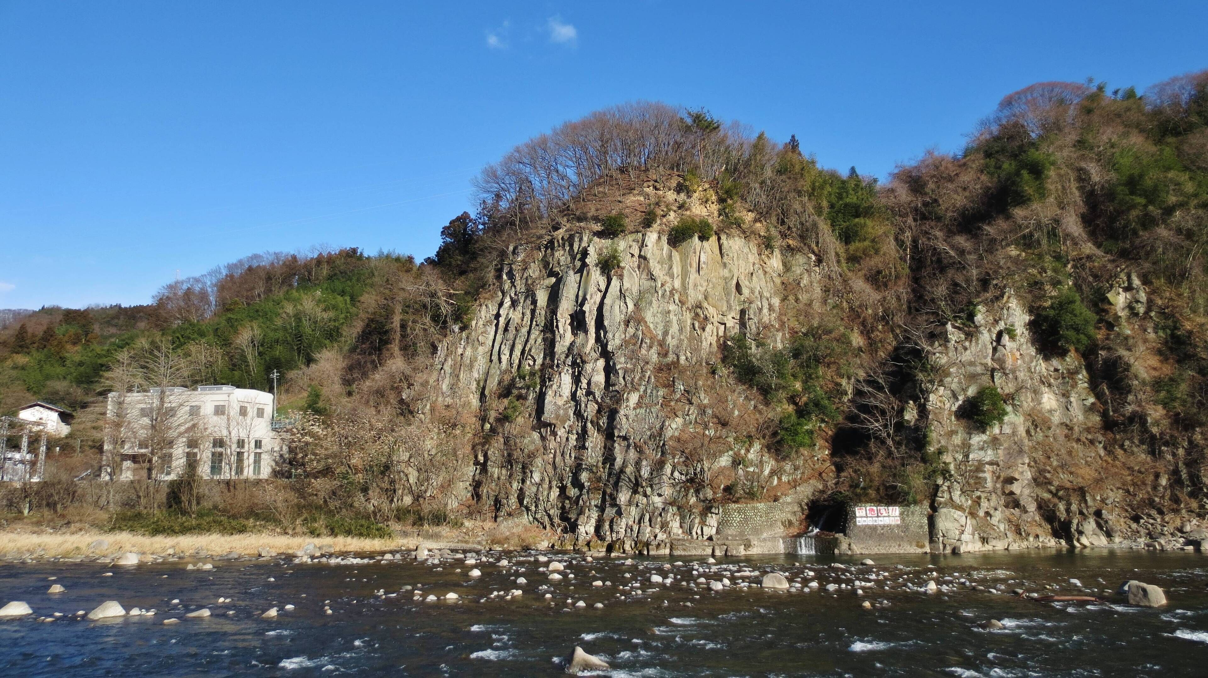 A river with rocks, a cliff with trees, and a building.