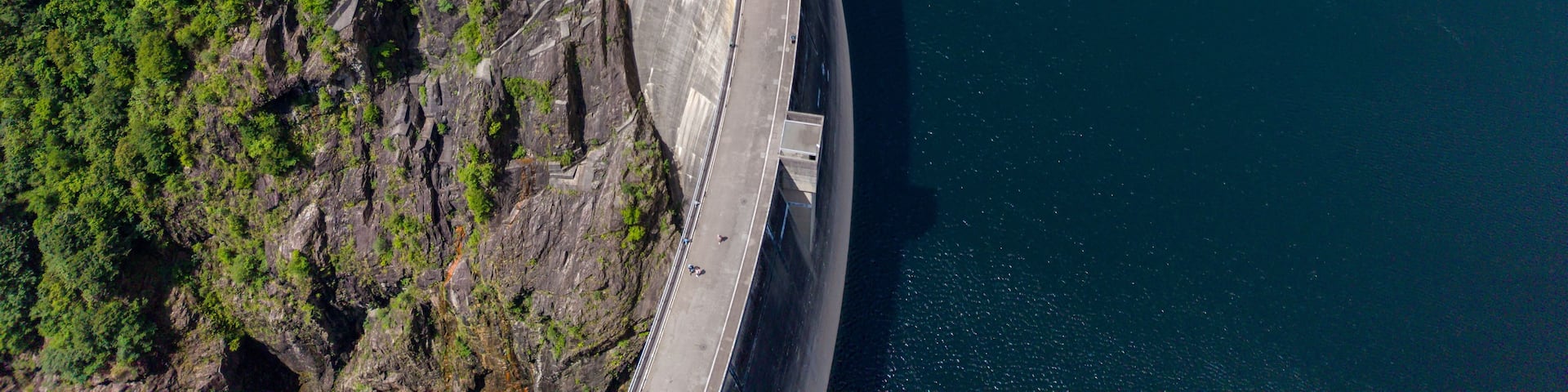 A large dam with a curved spillway, surrounded by dense forest and cliffs.