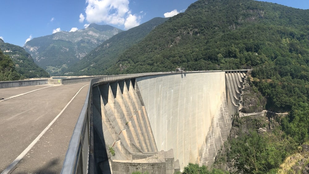 A large dam with a road running alongside it, surrounded by lush green mountains.