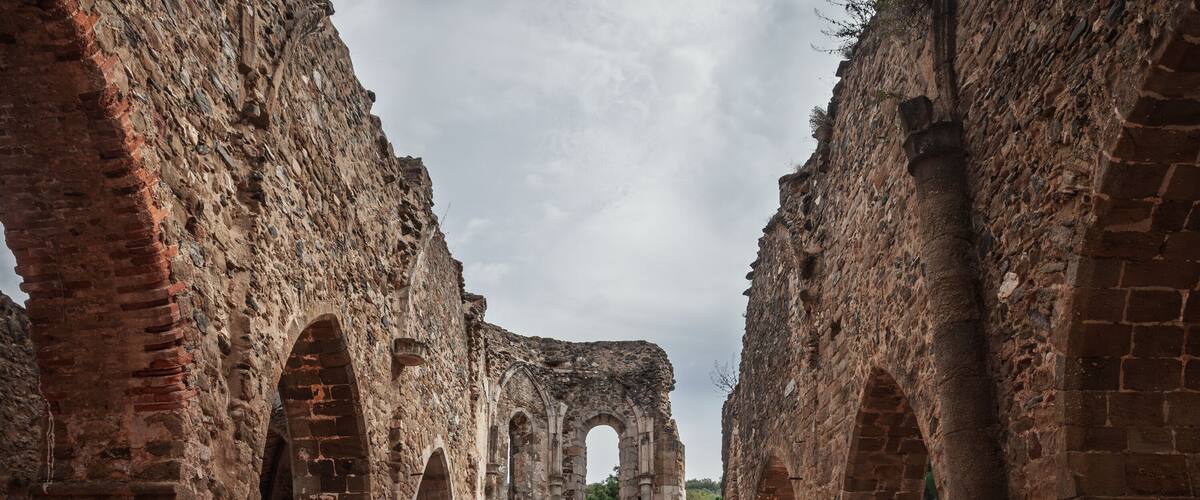 Ancient stone ruins with arches and columns, a paved area in the center, and a cloudy sky.