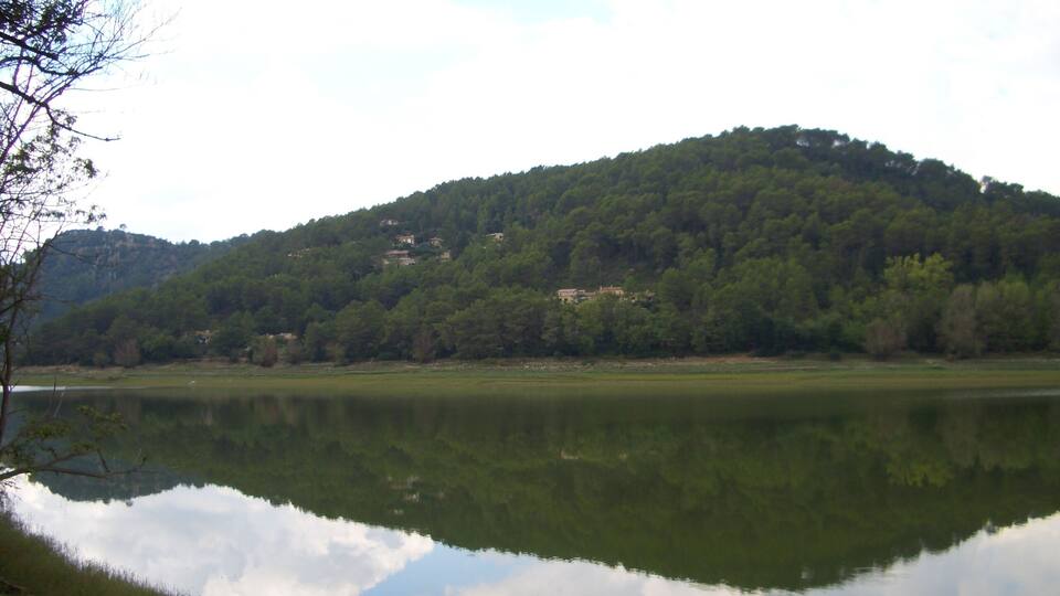 A calm lake with a reflection of the sky and trees, surrounded by a forested hillside and a few buildings in the distance.