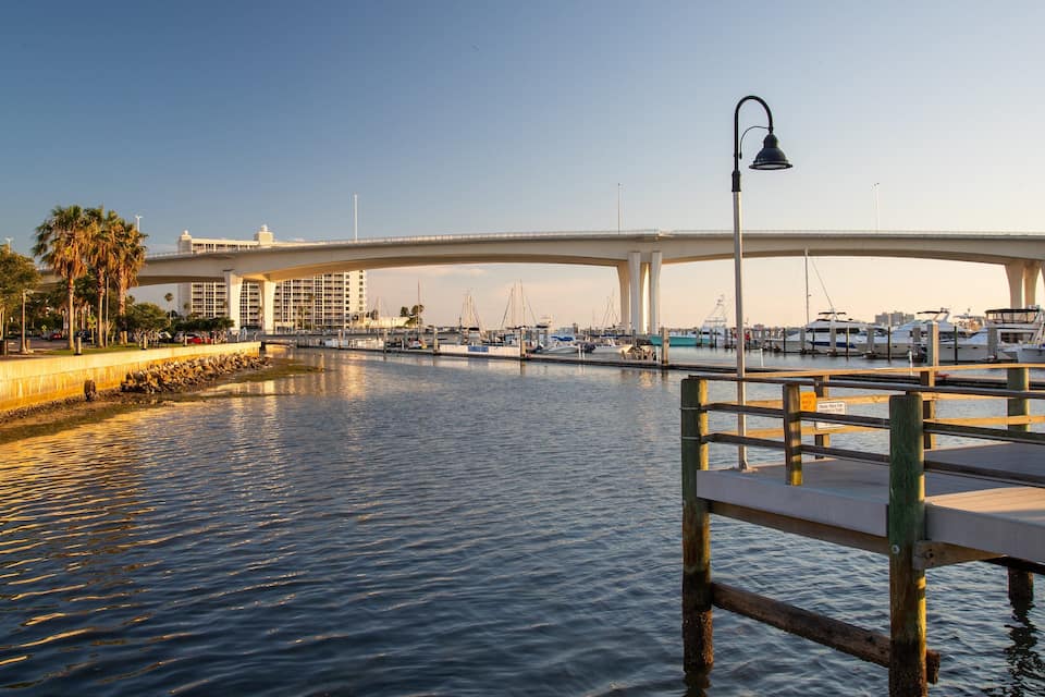 Uma marina com barcos atracados sob uma ponte, um calçadão de madeira e palmeiras na orla.