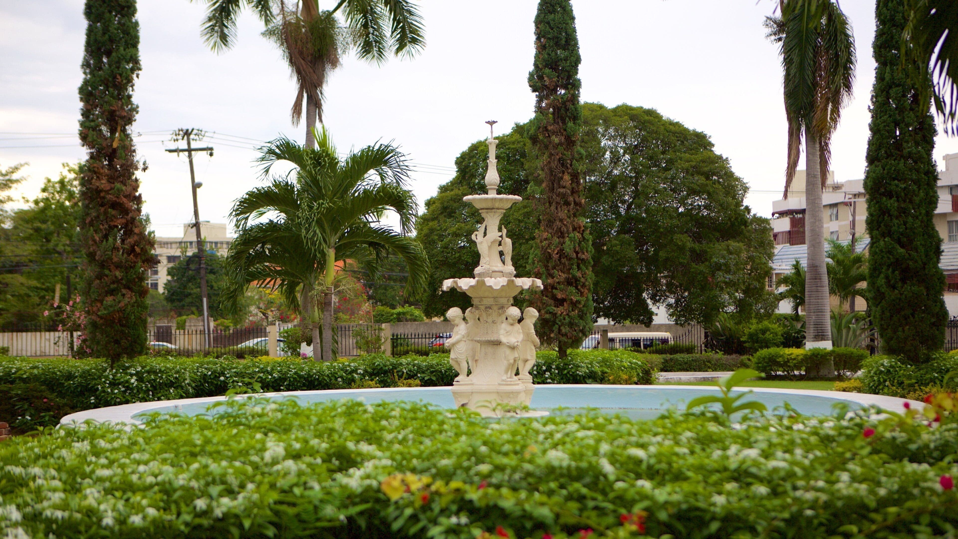 Uma fonte com estátuas em um jardim bem cuidado, com palmeiras e um edifício ao fundo.