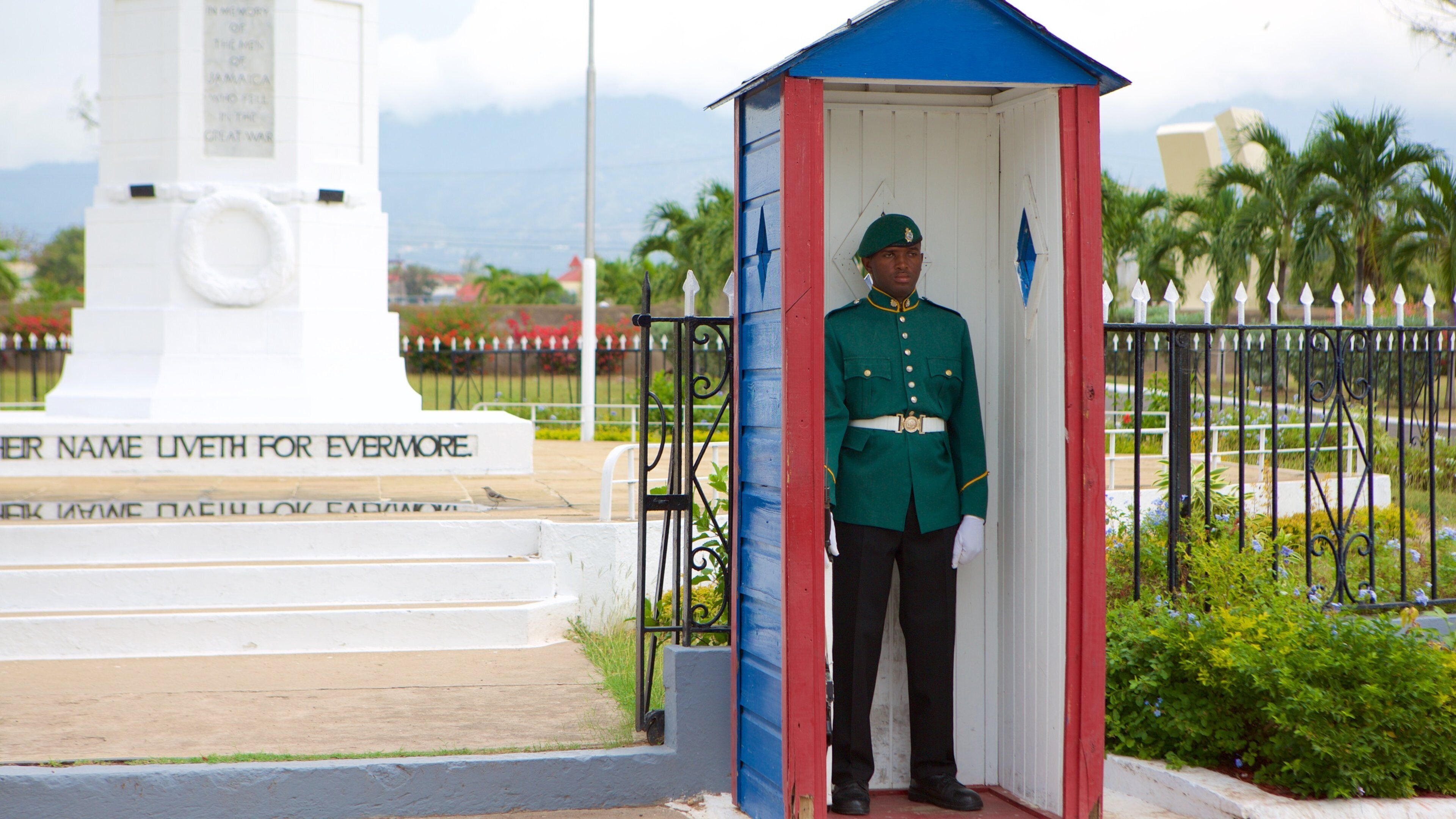 Un guardia uniformado de pie en un pequeño quiosco de fachada blanca, roja y azul. Al fondo, se aprecia un monumento blanco con inscripciones y vista al océano.