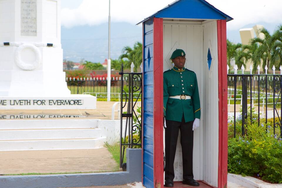 Un guardia uniformado de pie en un pequeño quiosco de fachada blanca, roja y azul. Al fondo, se aprecia un monumento blanco con inscripciones y vista al océano.