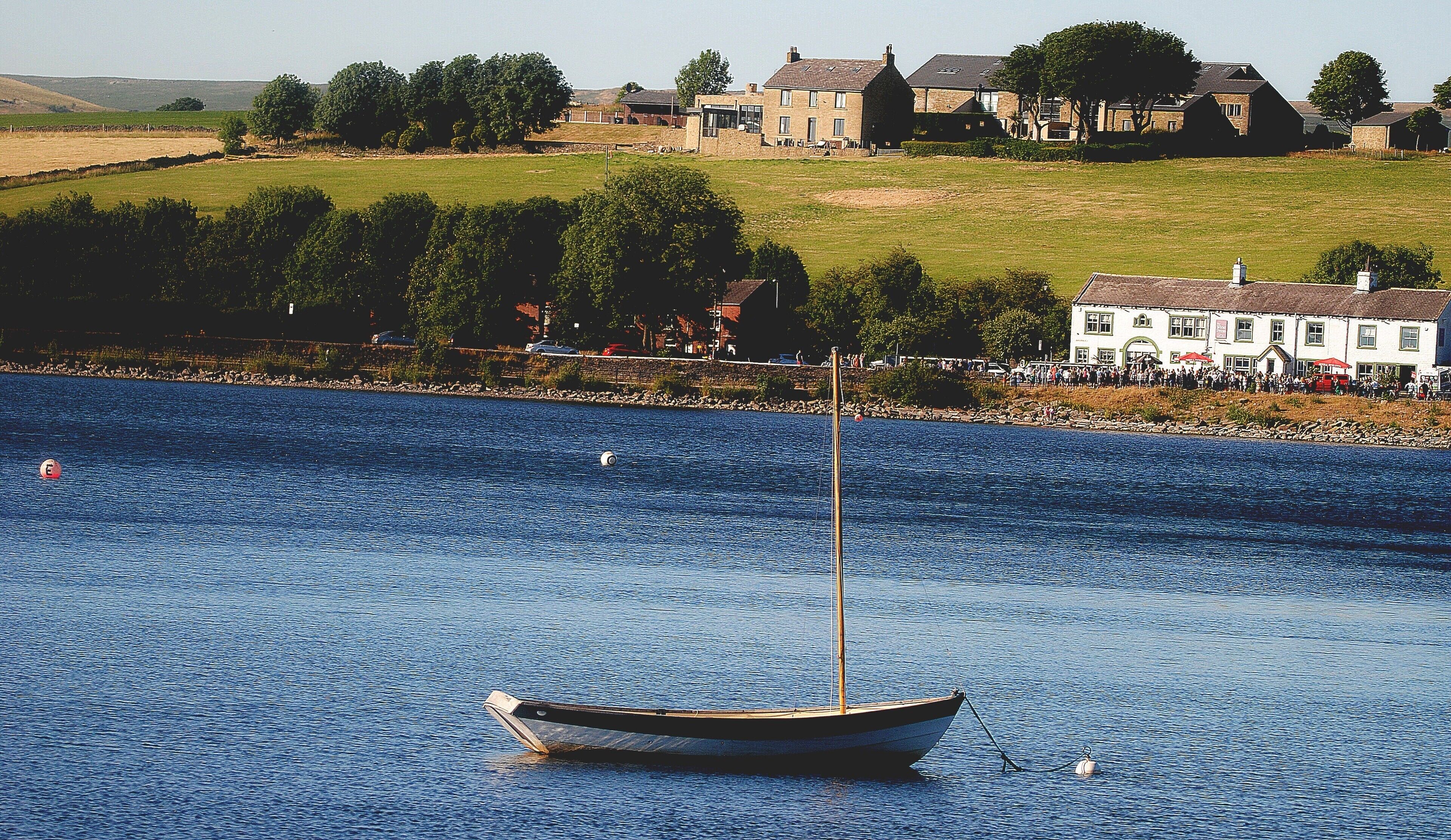 A calm body of water with a small sailboat anchored near the shore. In the background, there are houses and a grassy hill.