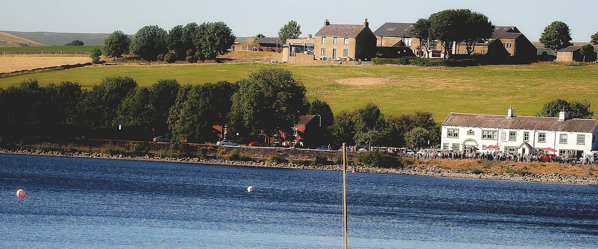 A calm body of water with a small sailboat anchored near the shore. In the background, there are houses and a grassy hill.