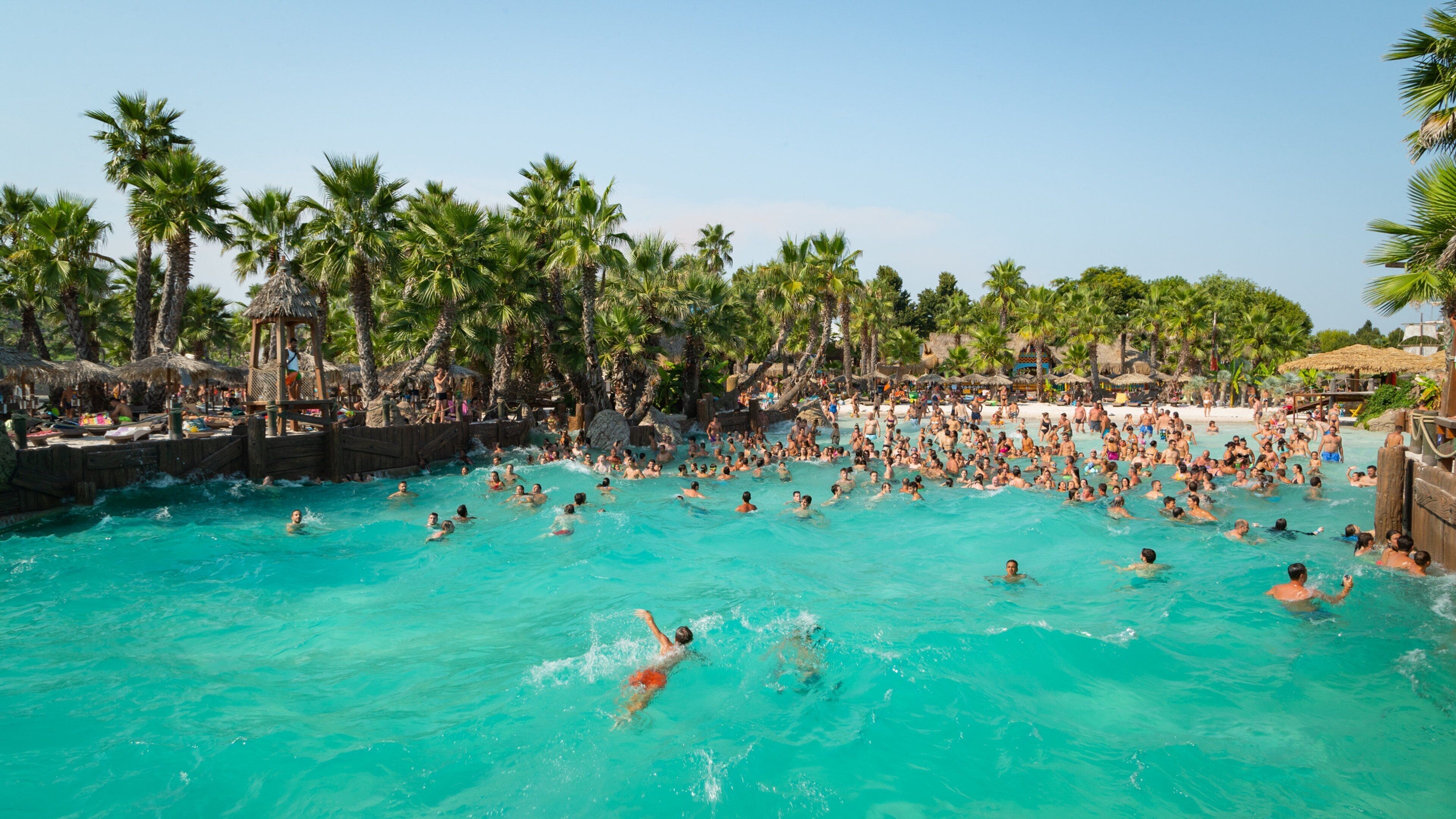 Una piscina concurrida de agua turquesa cristalina, rodeada de palmeras y estructuras con techo de paja.