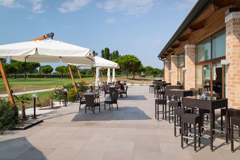 Een terras buiten met tafels en stoelen, een witte parasol en uitzicht op een golfbaan.