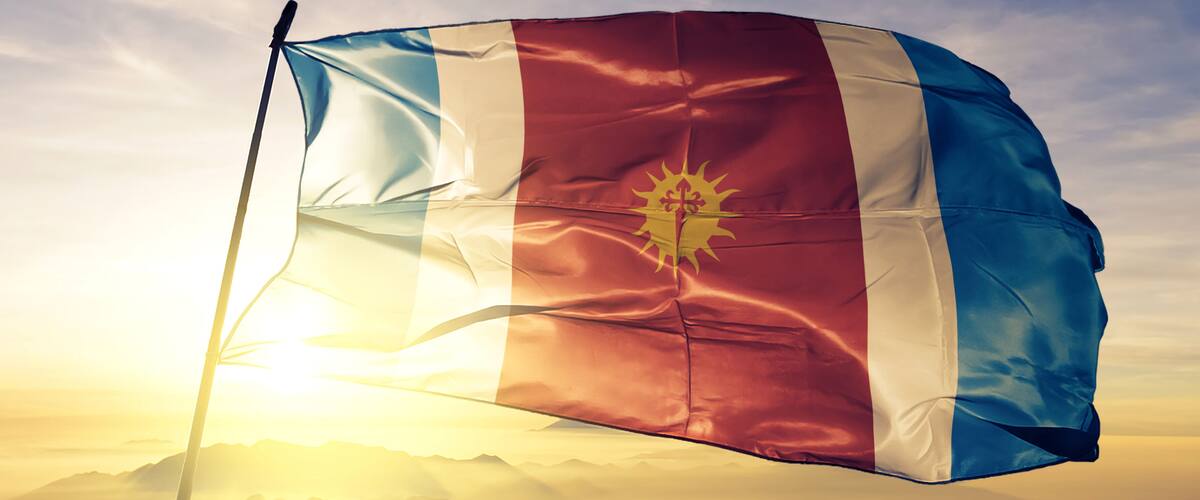 A flag waving against a clear sky with a sunlit mountain range in the background.