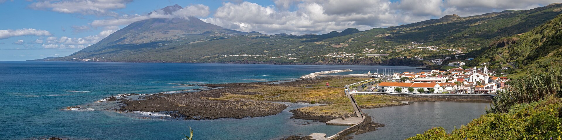 Uma vila costeira com uma montanha ao fundo, costa rochosa e vegetação exuberante.