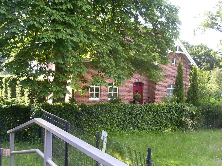 A red brick house with a white door, surrounded by lush greenery and a metal fence.