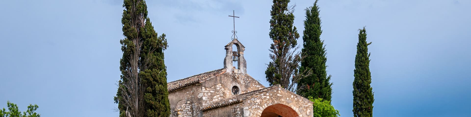 A stone church with a bell tower, surrounded by cypress trees and rocky terrain.