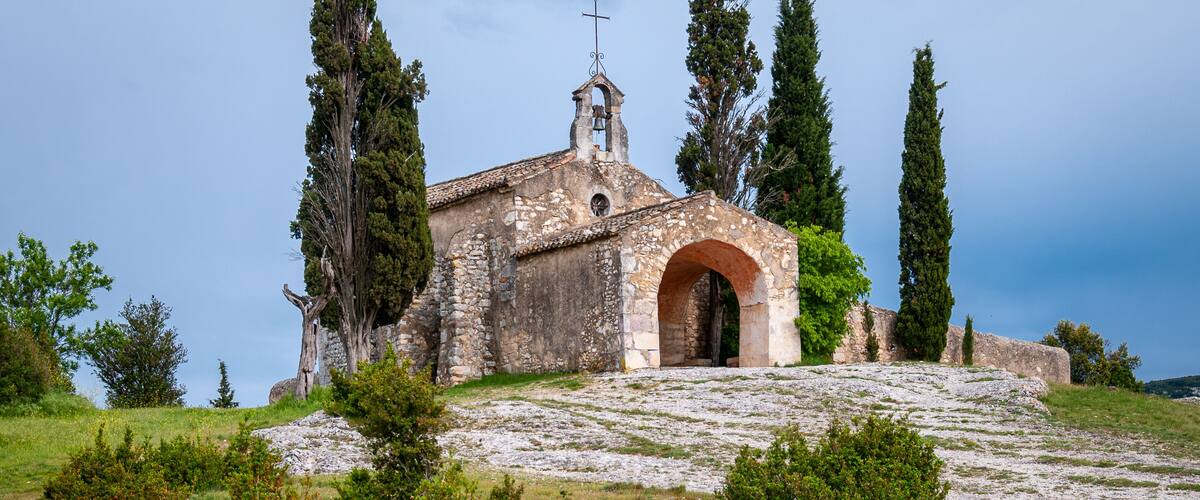 A stone church with a bell tower, surrounded by cypress trees and rocky terrain.