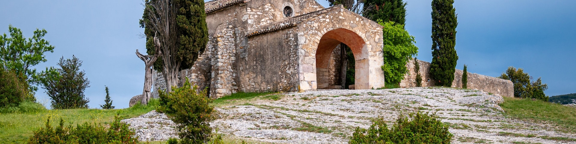A stone church with a bell tower, surrounded by cypress trees and rocky terrain.