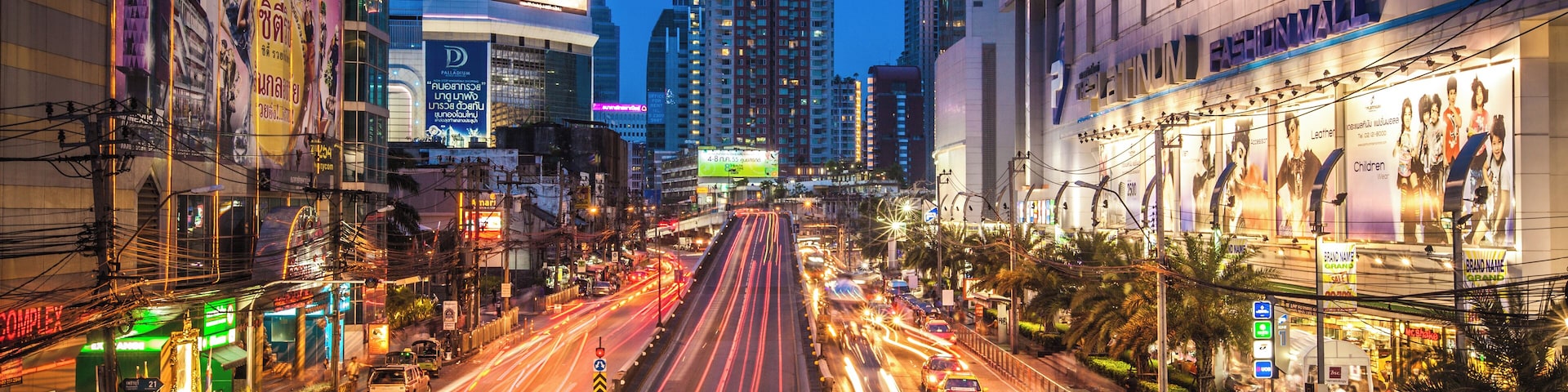 Uma rua urbana movimentada ao entardecer, com outdoors iluminados e edifícios altos.