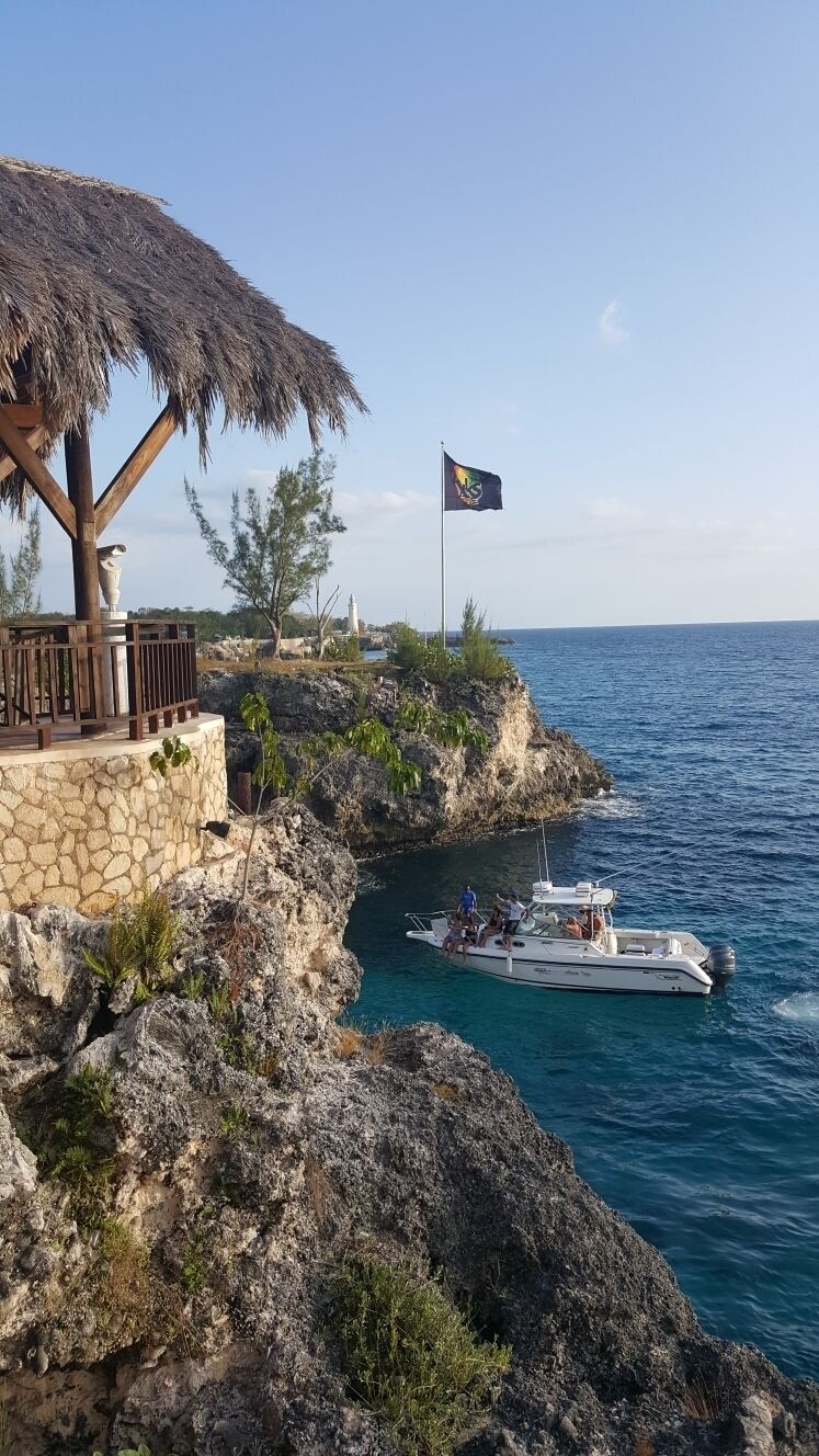 A coastal scene with a thatched-roof structure, a flag, rocky cliffs, and a boat in the water.