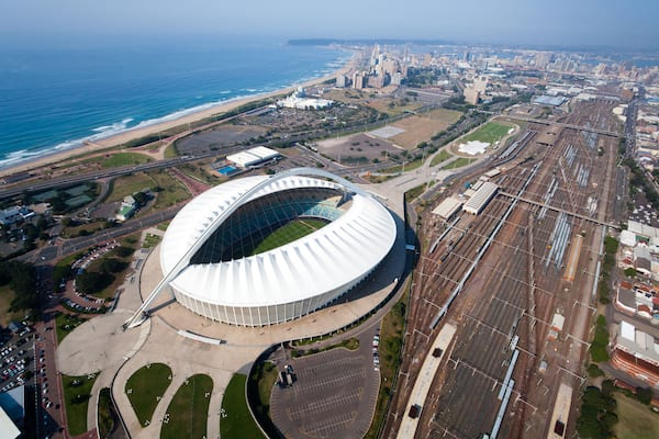 Un stade doté d’un toit blanc remarquable, un paysage urbain côtier avec une plage, et un vaste réseau ferroviaire.