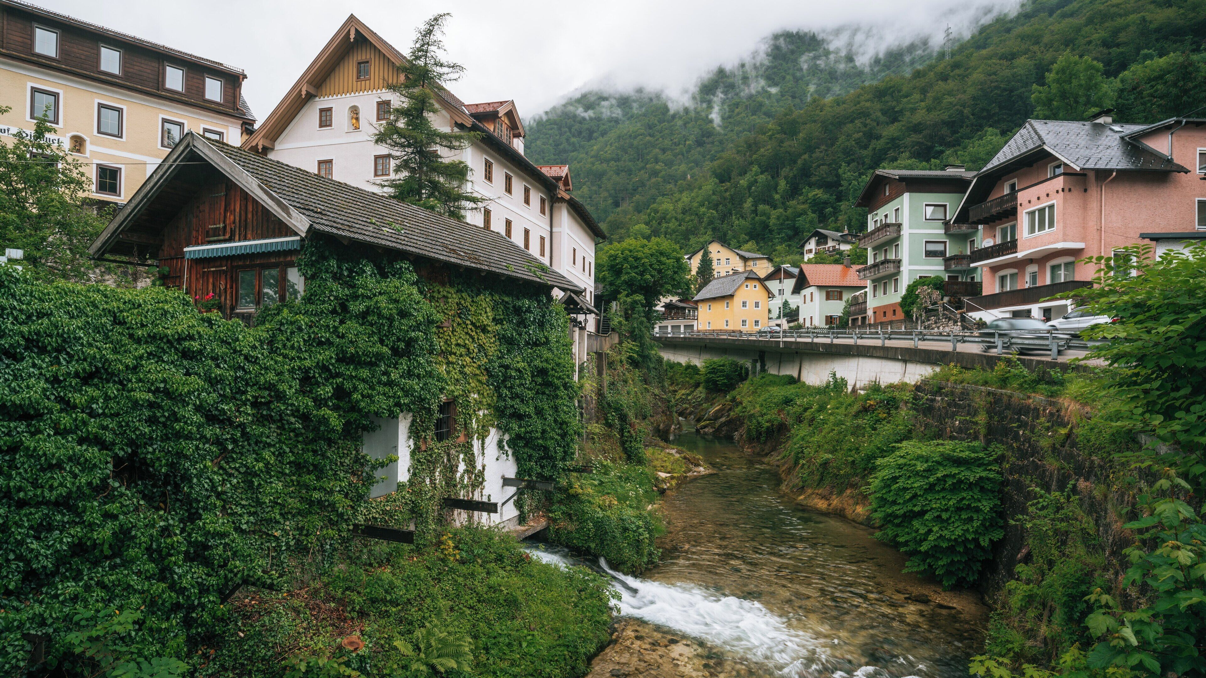 Un río serpentea por un pueblo con edificios a ambos lados, rodeado de exuberante vegetación y colinas al fondo.