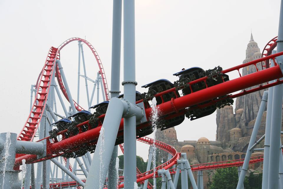 A roller coaster with red tracks and cars, set against a backdrop of a large, historic building with domed structures.
