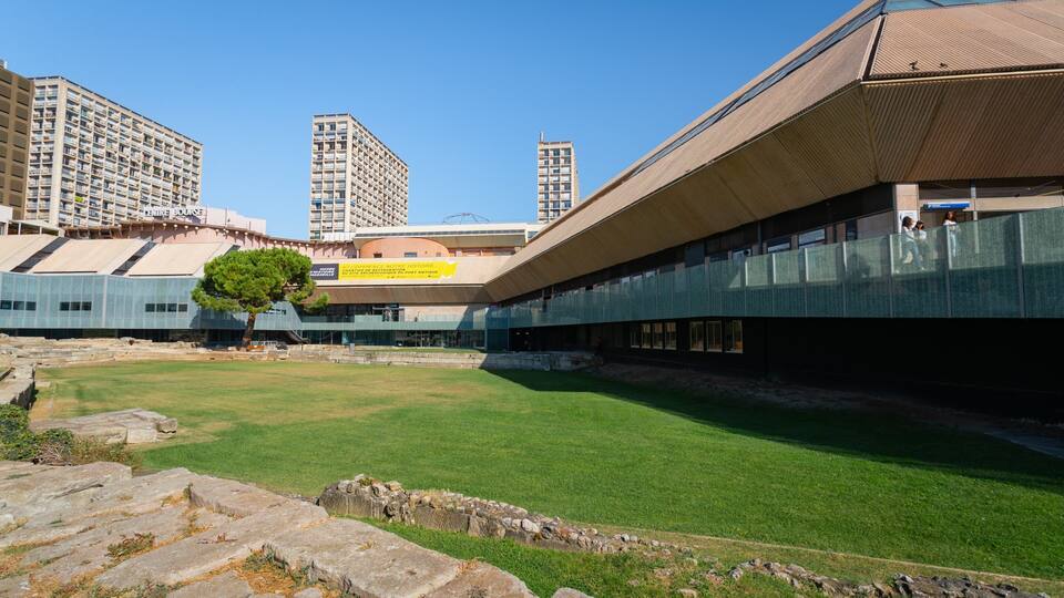 A modern building with a glass facade, a grassy courtyard, and ancient ruins in the foreground.