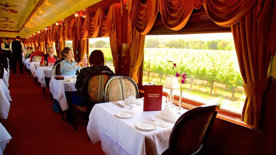 Una carrozza ristorante con tavoli apparecchiati per un pasto, ampie finestre con tende e vista sui vigneti.