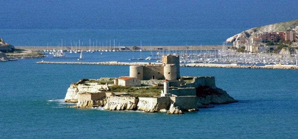 A coastal fortress with towers and a lighthouse, surrounded by calm sea and a marina with boats.