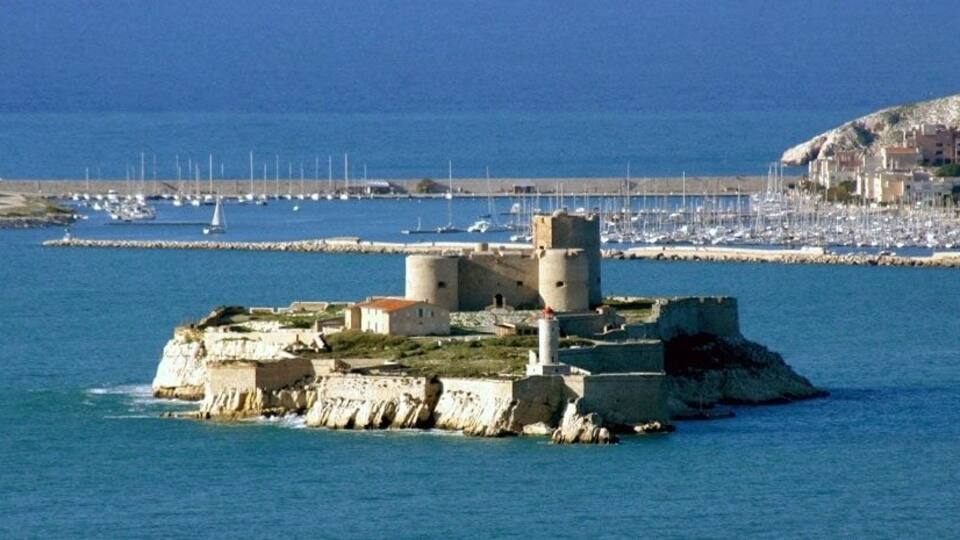 A coastal fortress with towers and a lighthouse, surrounded by calm sea and a marina with boats.