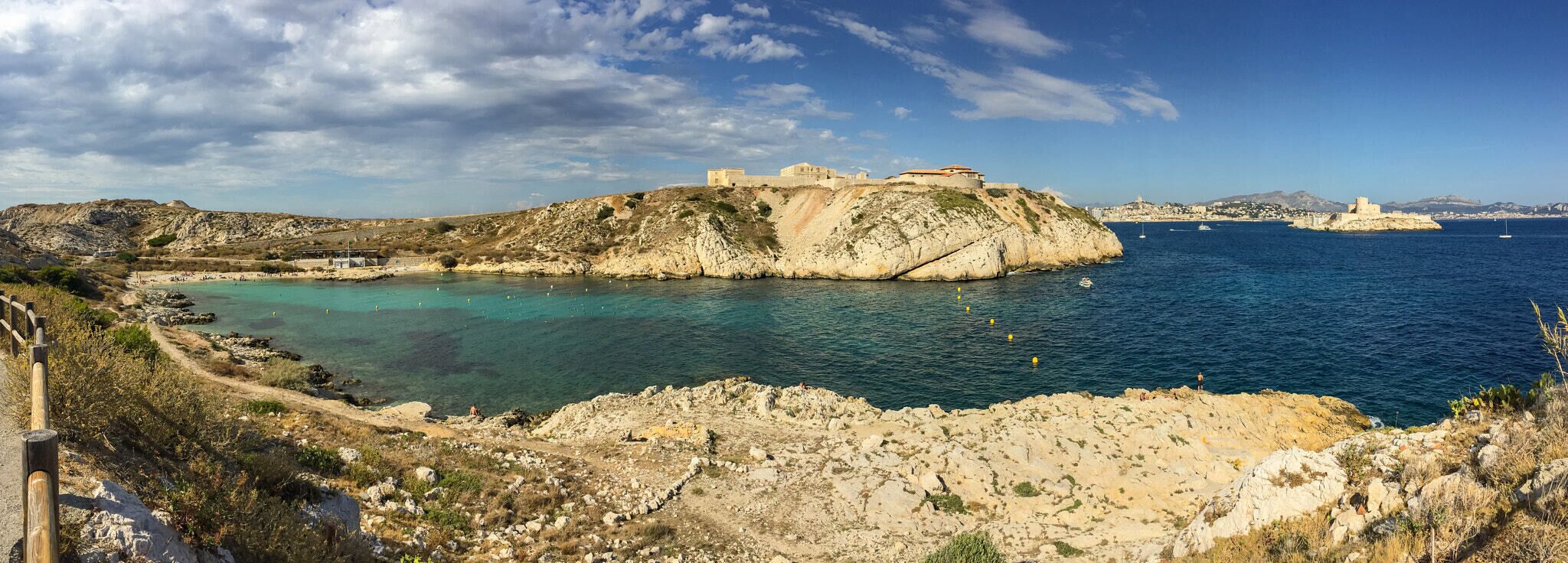 A coastal landscape with rocky terrain, a fortified structure on a hill, and a clear blue sea with boats.