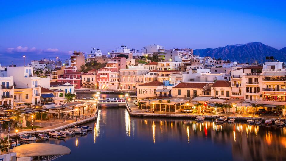 A coastal town at dusk with a marina, buildings, and mountains in the background.