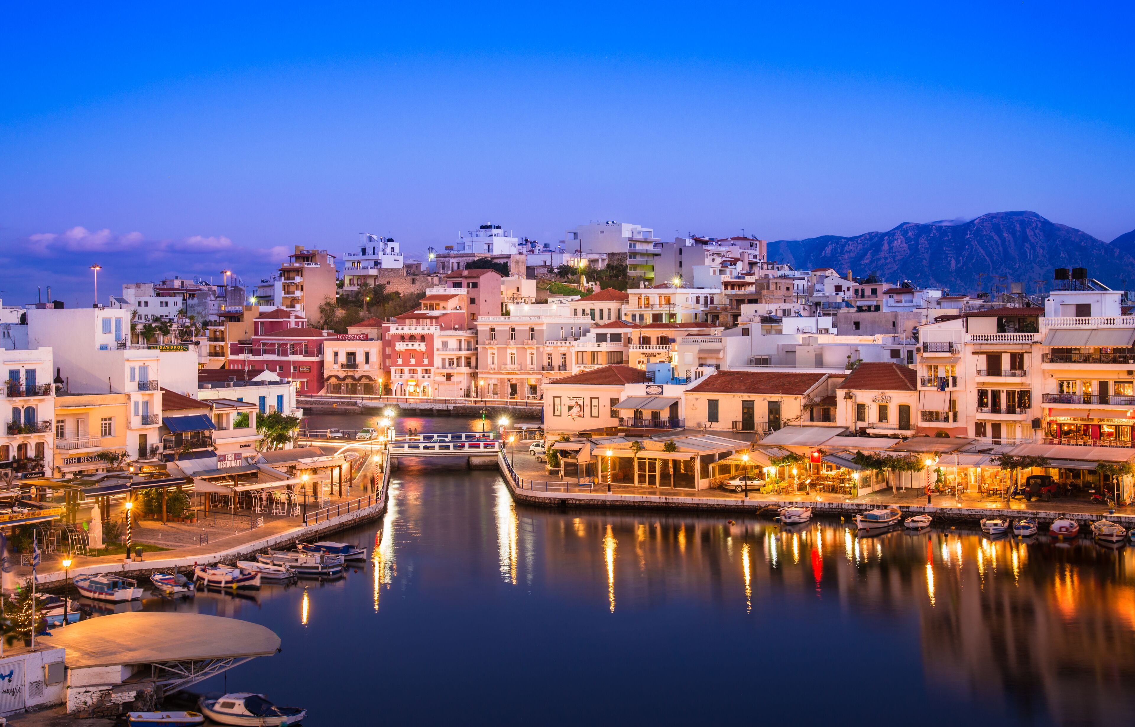 A coastal town at dusk with a marina, buildings, and mountains in the background.