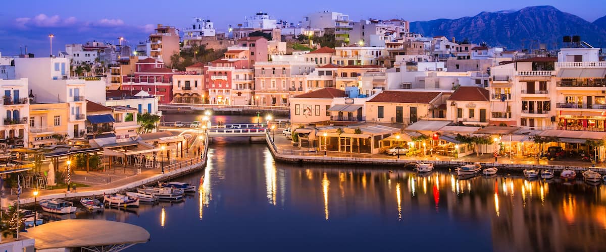 A coastal town at dusk with a marina, buildings, and mountains in the background.