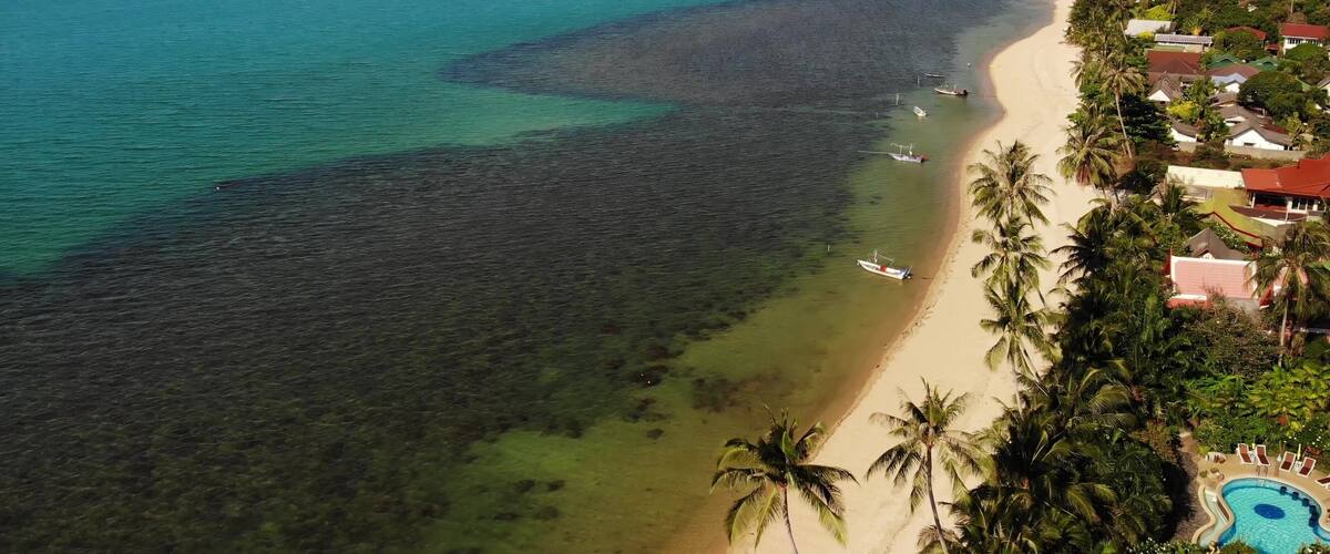 A coastal landscape with a clear turquoise sea, a sandy beach, and a resort area with palm trees and a swimming pool.