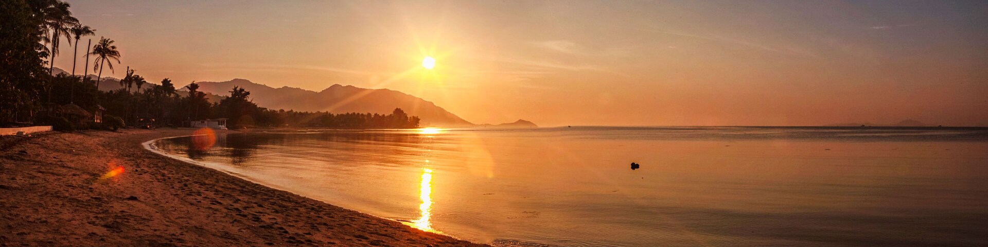 A beach at sunset with a clear sky, palm trees, and mountains in the background.