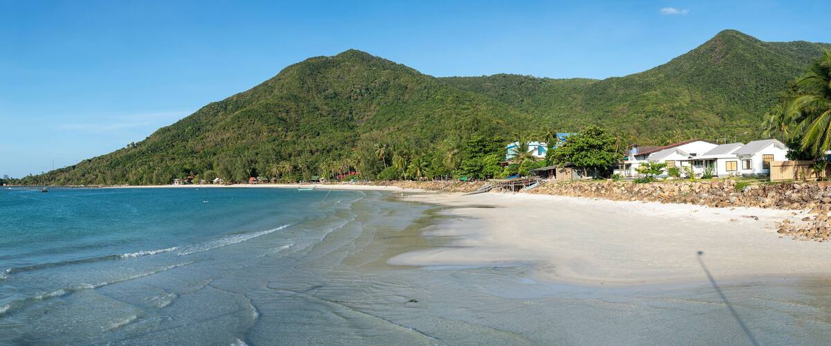 A beach with clear blue water, sandy shore, and lush green mountains in the background.