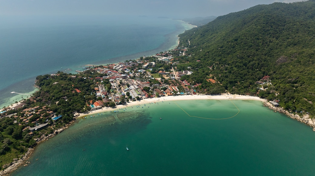 An aerial view of a coastal town with a beach, clear turquoise waters, and a mountainous backdrop.